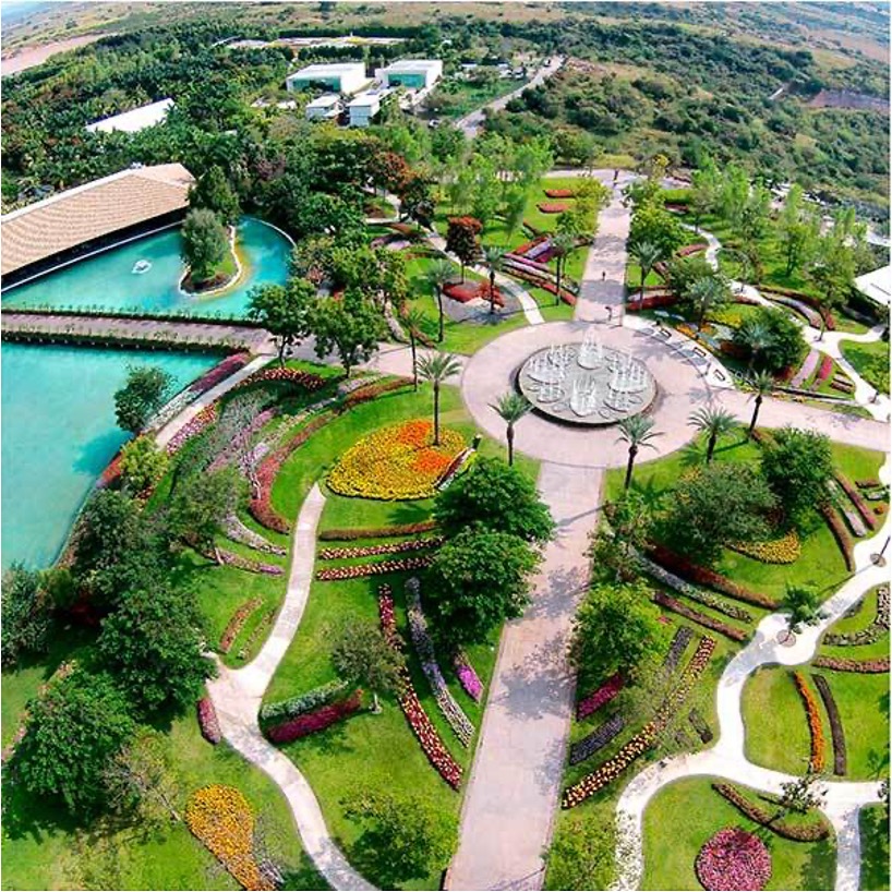 La imagen se muestra en formato panorámico y revela un extenso espacio de vegetación. En el centro de un amplio jardín se observa una fuente redonda en funcionamiento. Desde la fuente, se extienden cuatro pasillos que llevan a diferentes jardines. En la parte derecha, se puede ver un lago y un gran puente que lo cruza
La imagen muestra de manera panorámica una zona amplia con edificaciones. Se destacan cuatro edificios muy grandes rodeados de vegetación, árboles y albercas, lo que le da una apariencia de zona residencial. A los lados se pueden ver más edificaciones
