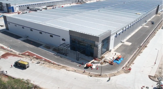 La imagen se muestra en formato panorámico y revela una construcción muy amplia de color gris, que parece ser una fábrica. Se observa una entrada de vidrio grande y vehículos estacionados afuera. Detrás de esta explanada, hay otra construcción muy larga en la parte posterior
La imagen muestra de manera panorámica una zona amplia con edificaciones. Se destacan cuatro edificios muy grandes rodeados de vegetación, árboles y albercas, lo que le da una apariencia de zona residencial. A los lados se pueden ver más edificaciones